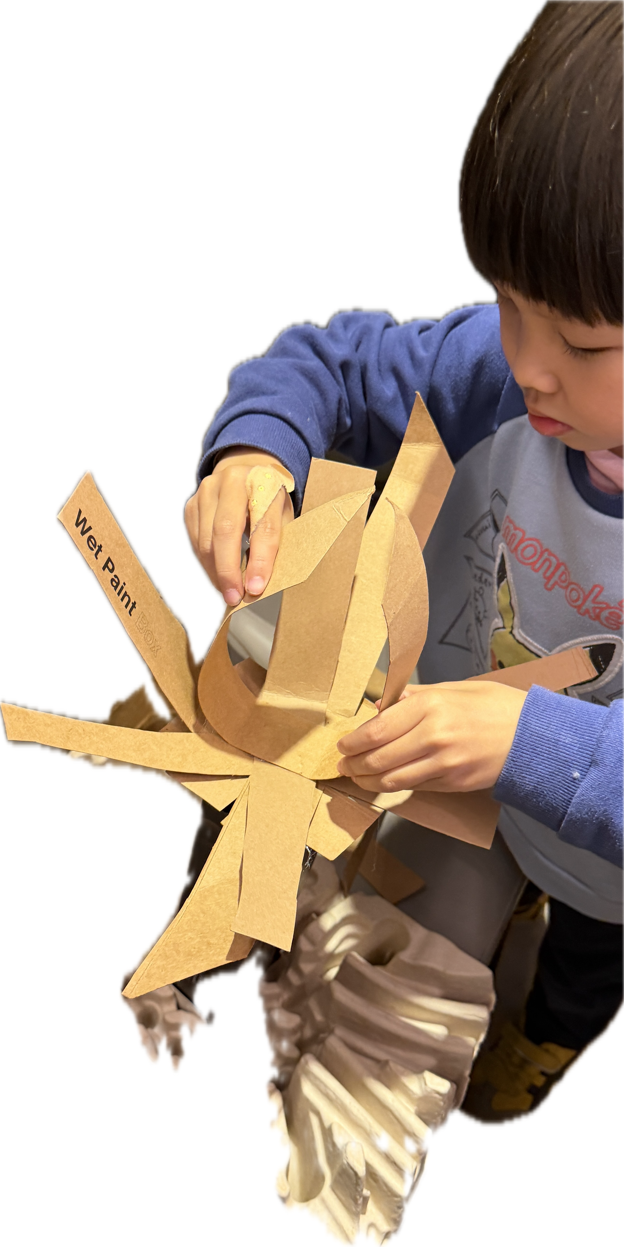 A young boy is working on a craft project with a large paper butterfly made from brown paper with the words "Wet Paint" on one of its wings. He is wearing a blue and gray shirt with cartoon characters, and a bandage on his finger.