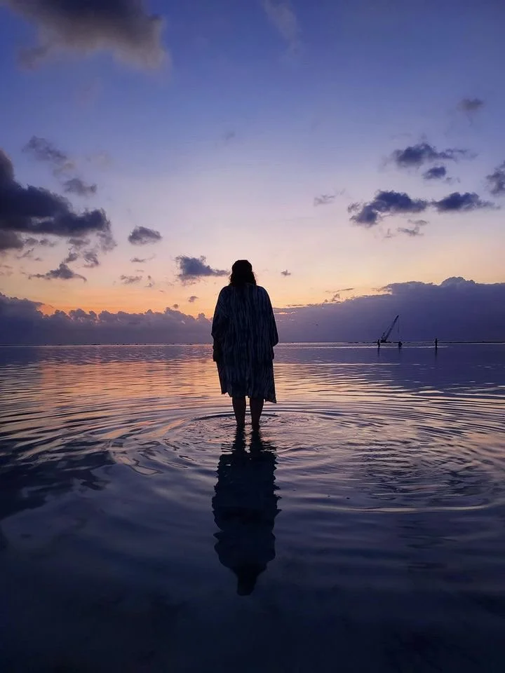 Jess standing in shallow ocean water at sunset, silhouetted against a purple and orange sky, looking towards the horizon.