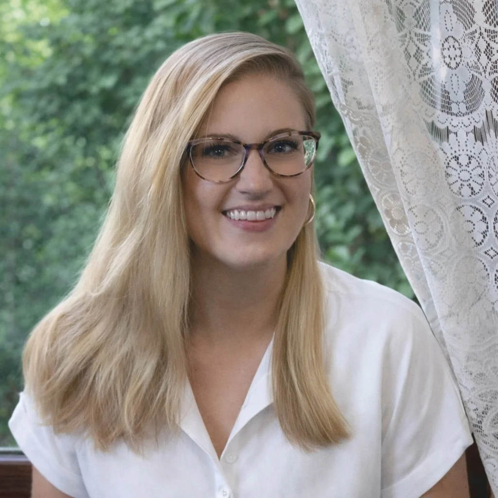 A young woman with blonde hair and glasses smiling, sitting near a window with lace curtains, with greenery outside.