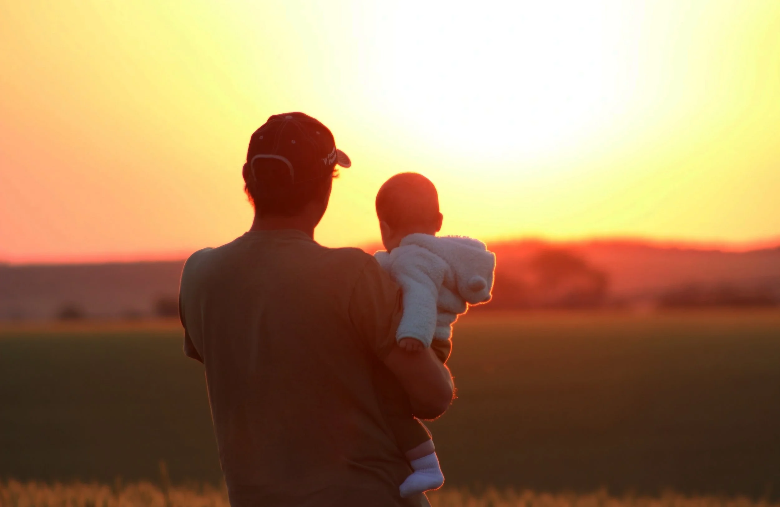 a man with a hat, holding his infant daughter with his back facing the foreground watching a beautiful sunrise