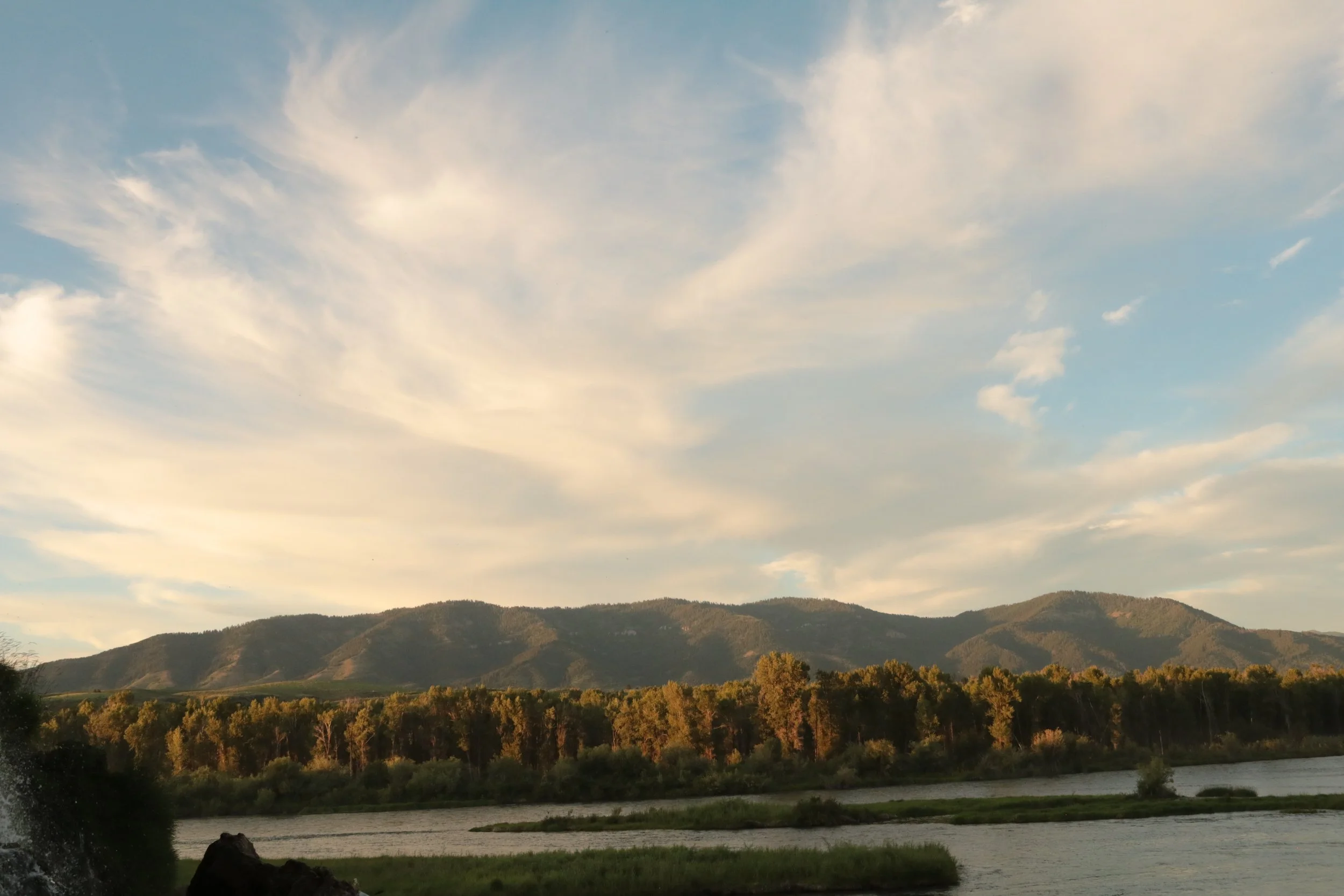 A scenic landscape featuring a river in the foreground, surrounded by trees, with rolling hills and mountains in the background under a partly cloudy sky.