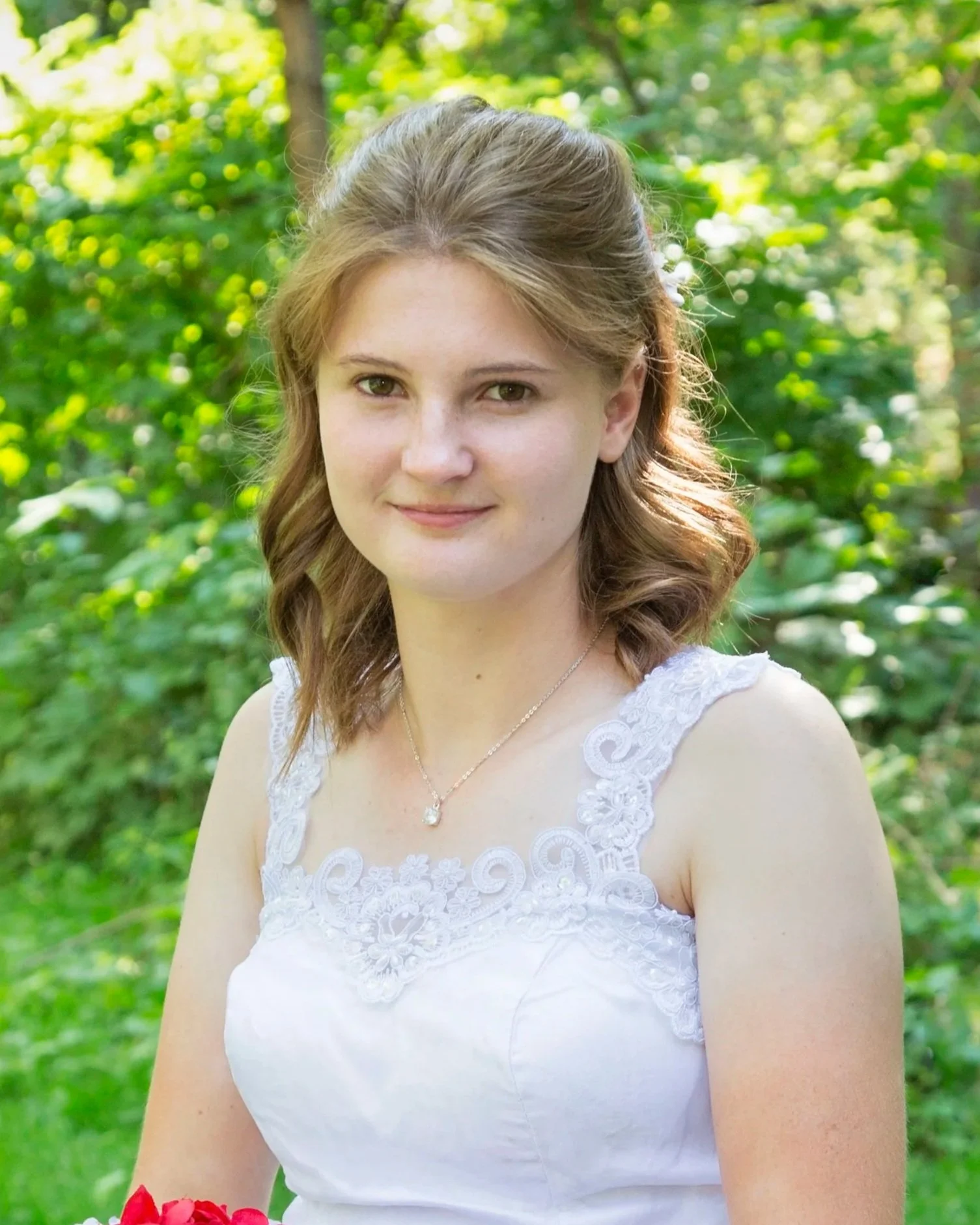 Portrait of a young woman in a white dress with lace details, standing outdoors amidst green foliage, smiling gently at the camera.