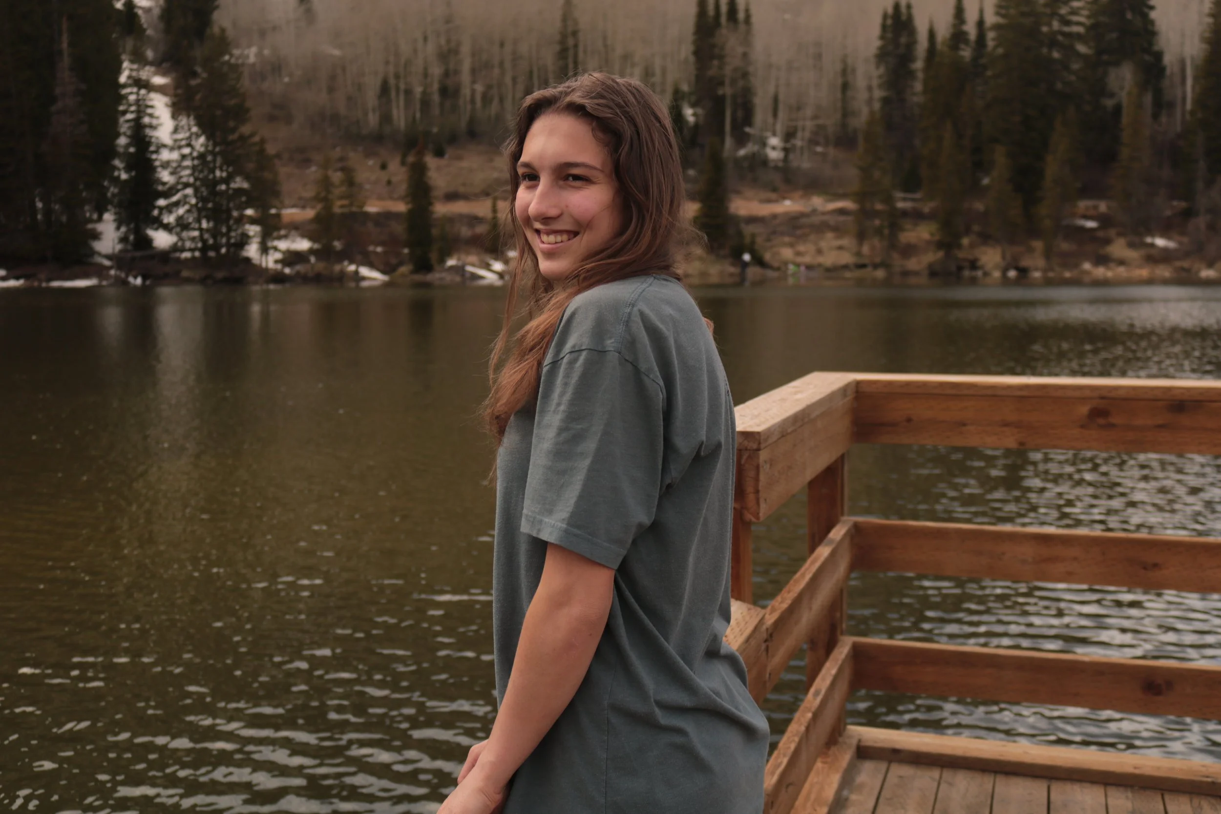 A young woman with long brown hair smiling and standing on a wooden dock by a lake, with trees and a hillside in the background.