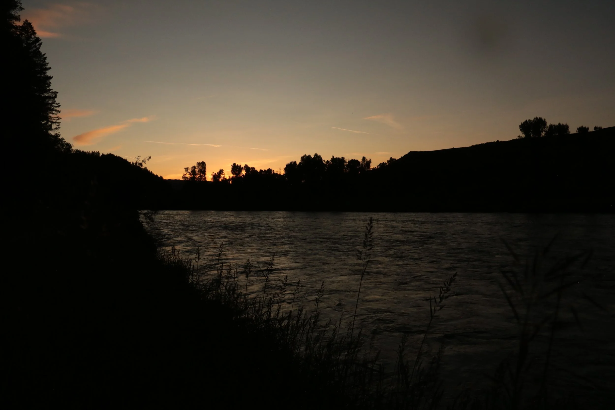 Sunset over a river with silhouetted trees and hills, with a dark sky transitioning from light near the horizon.