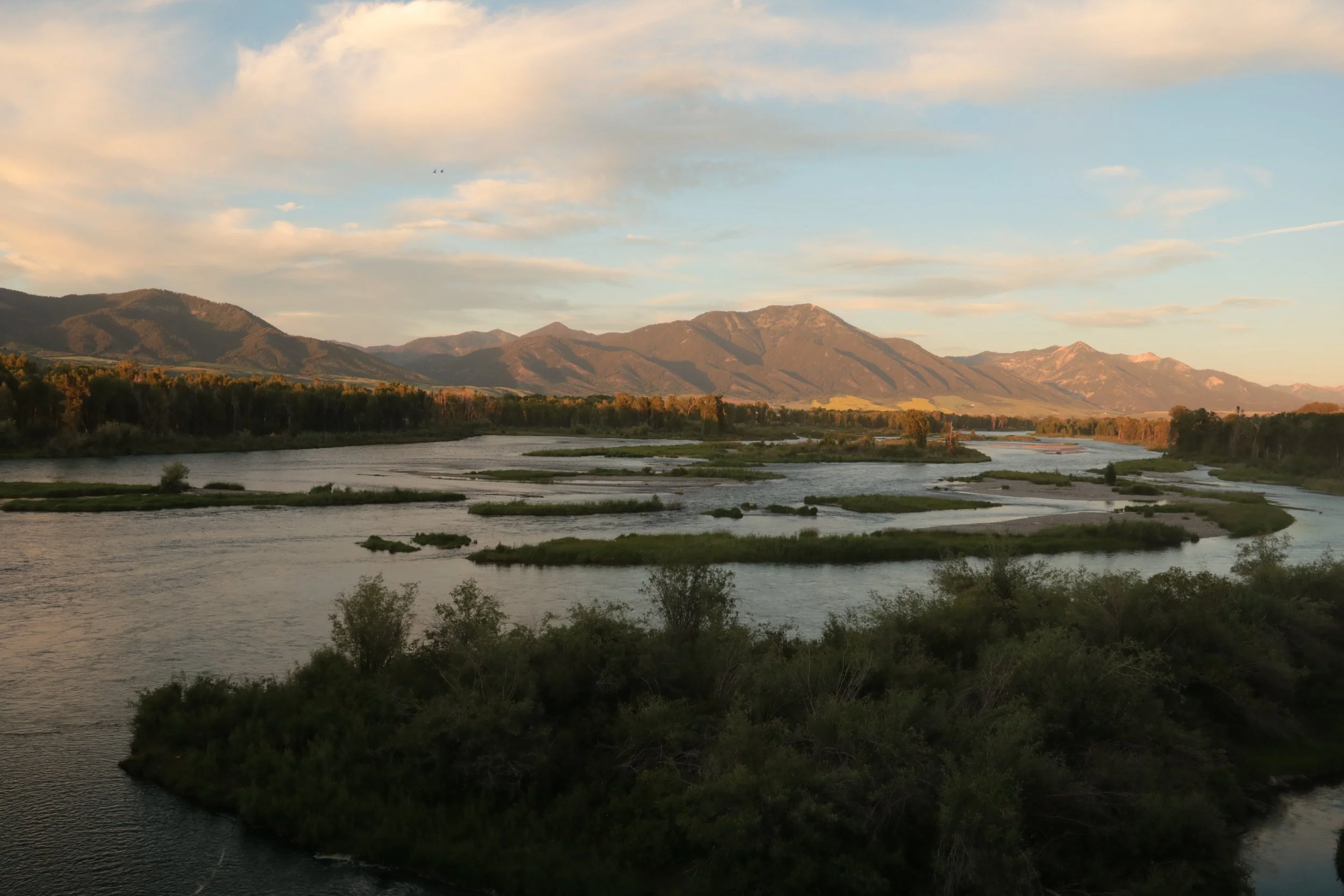 A landscape with a river flowing through green vegetation with mountains in the background and a partly cloudy sky.
