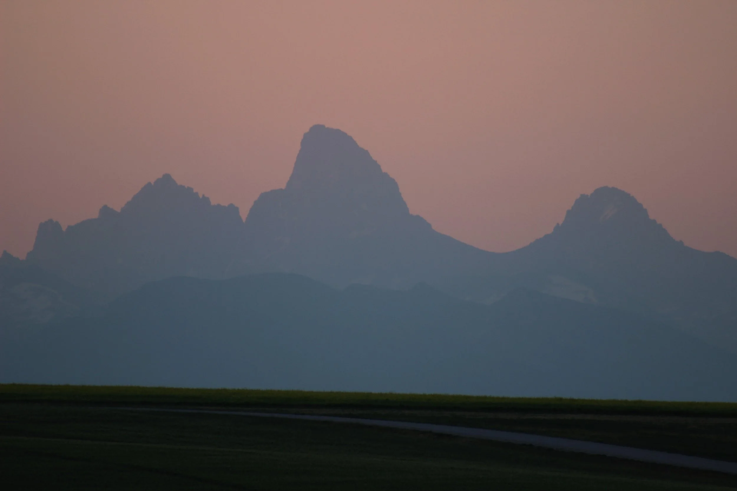 a light blue silhouette of the Teton mountain  peaks set against a pink sunrise sky with a darker foreground