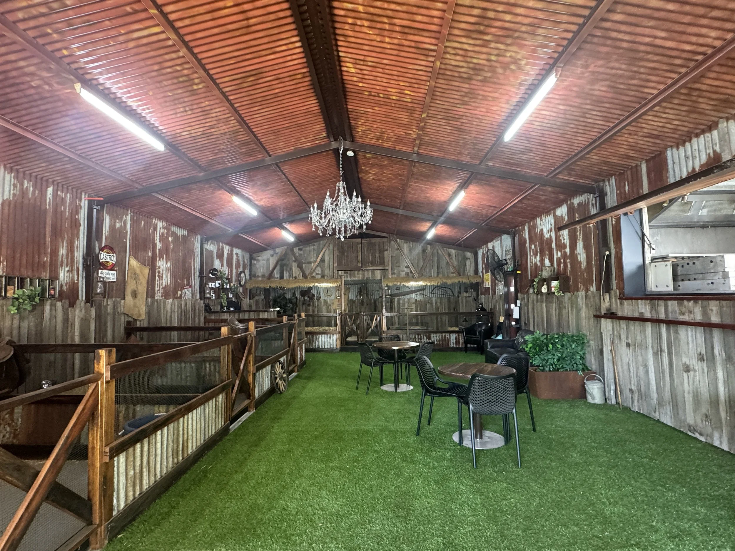 A rustic indoor space with wooden walls and ceiling, decorated with a chandelier, artificial turf flooring, and black chairs around tables.