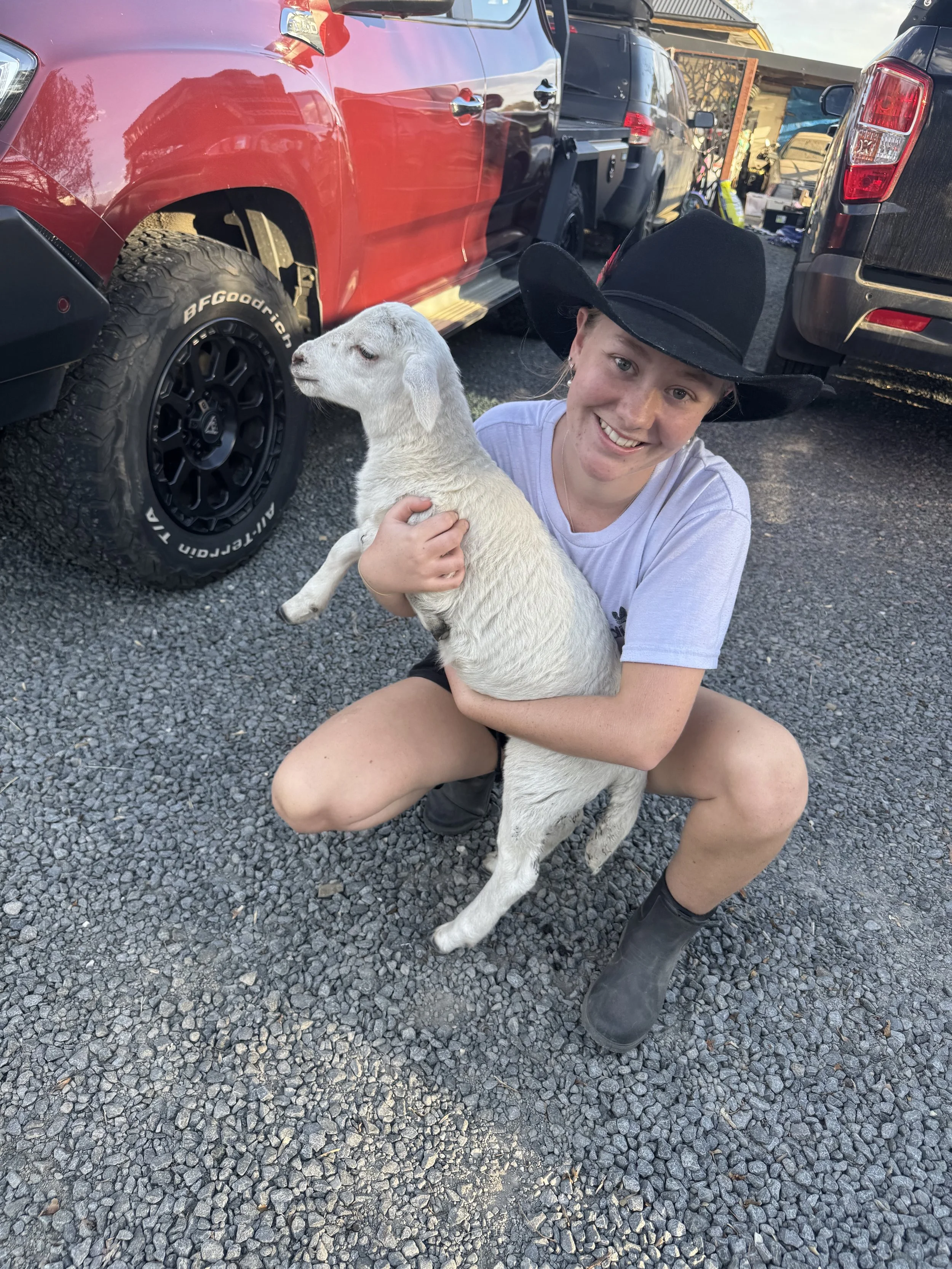 A smiling woman wearing a black cowboy hat, gray t-shirt, black shorts, and rubber boots is crouching on gravel and holding a young white goat. The goat is standing on its hind legs, leaning into the woman's embrace, with a neutral expression. Several vehicles are parked on gravel behind them, including a red vehicle with large tires, and a black vehicle with a roof rack. A suburban house and a wooden fence are visible in the background.