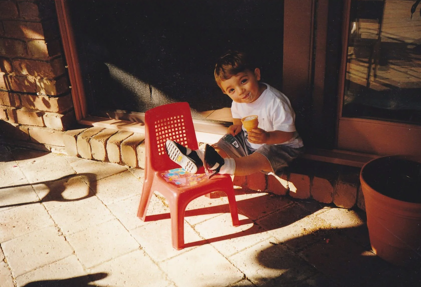 A young boy sitting outside near a brick fireplace, holding an ice cream cone, with a red plastic chair in front of him and a large potted plant nearby.