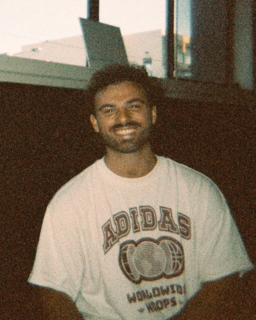 A man with dark curly hair and a beard, smiling, wearing a white Adidas t-shirt with a graphic design and text, sitting indoors near a window with a laptop on a table in the background.