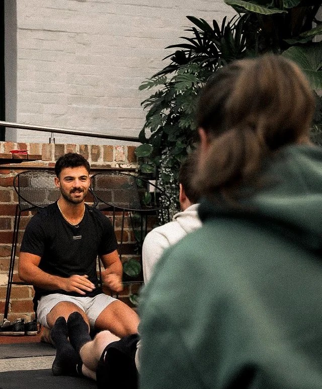 A group of four young people sitting on the floor, engaged in a conversation or activity with one person speaking. The setting appears to be indoors with a brick wall, greenery, and patio furniture in the background.