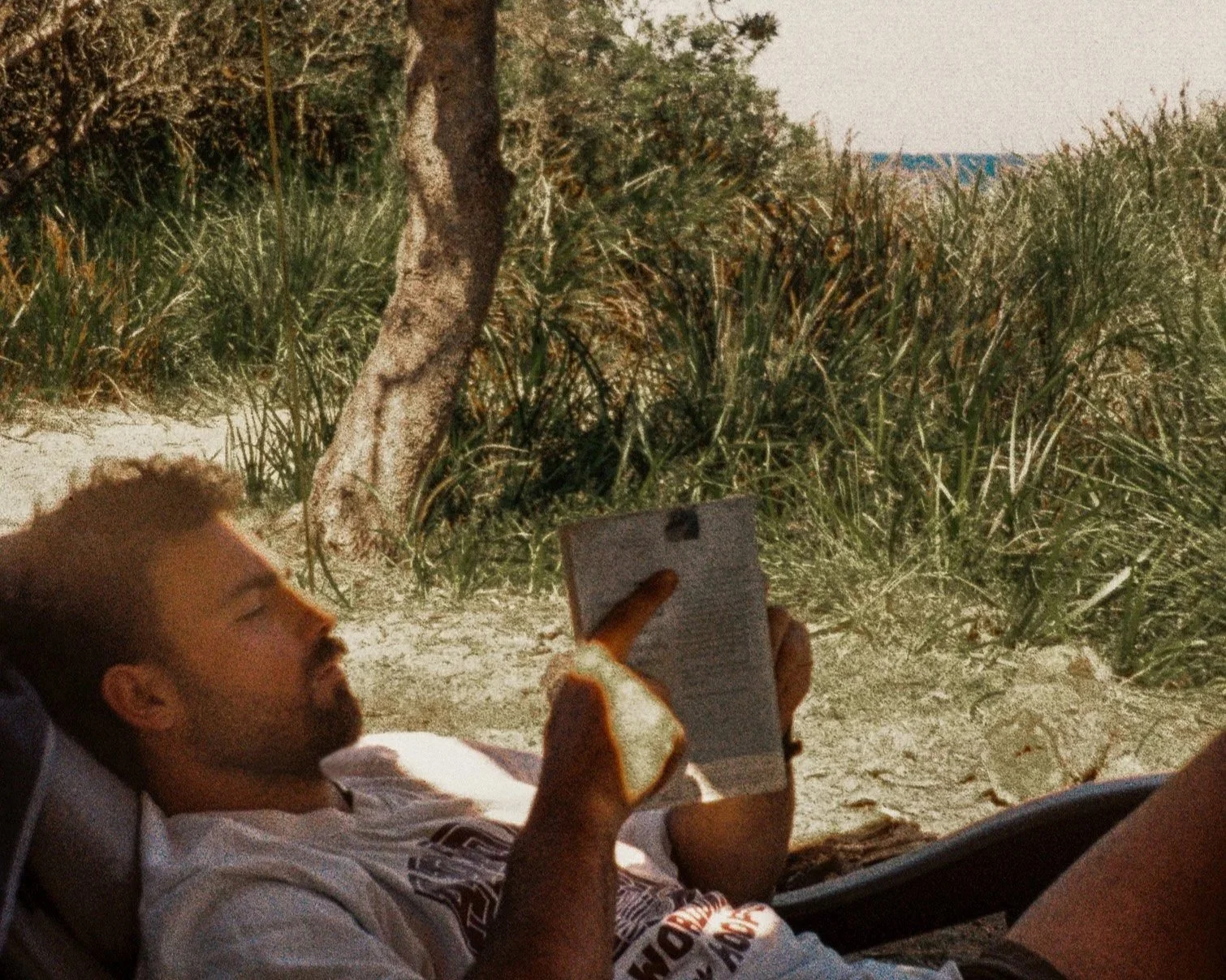 A man with short hair and a beard is lying on a chair outdoors, reading a book while holding a slice of bread, with a background of trees and vegetation.