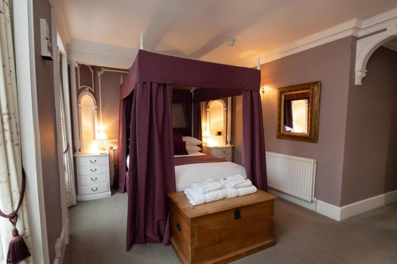 Bedroom with four-poster bed with purple drapes, wooden chest at foot of bed with folded towels, nightstands with lamps, framed mirror on wall, and neutral-colored curtains.
