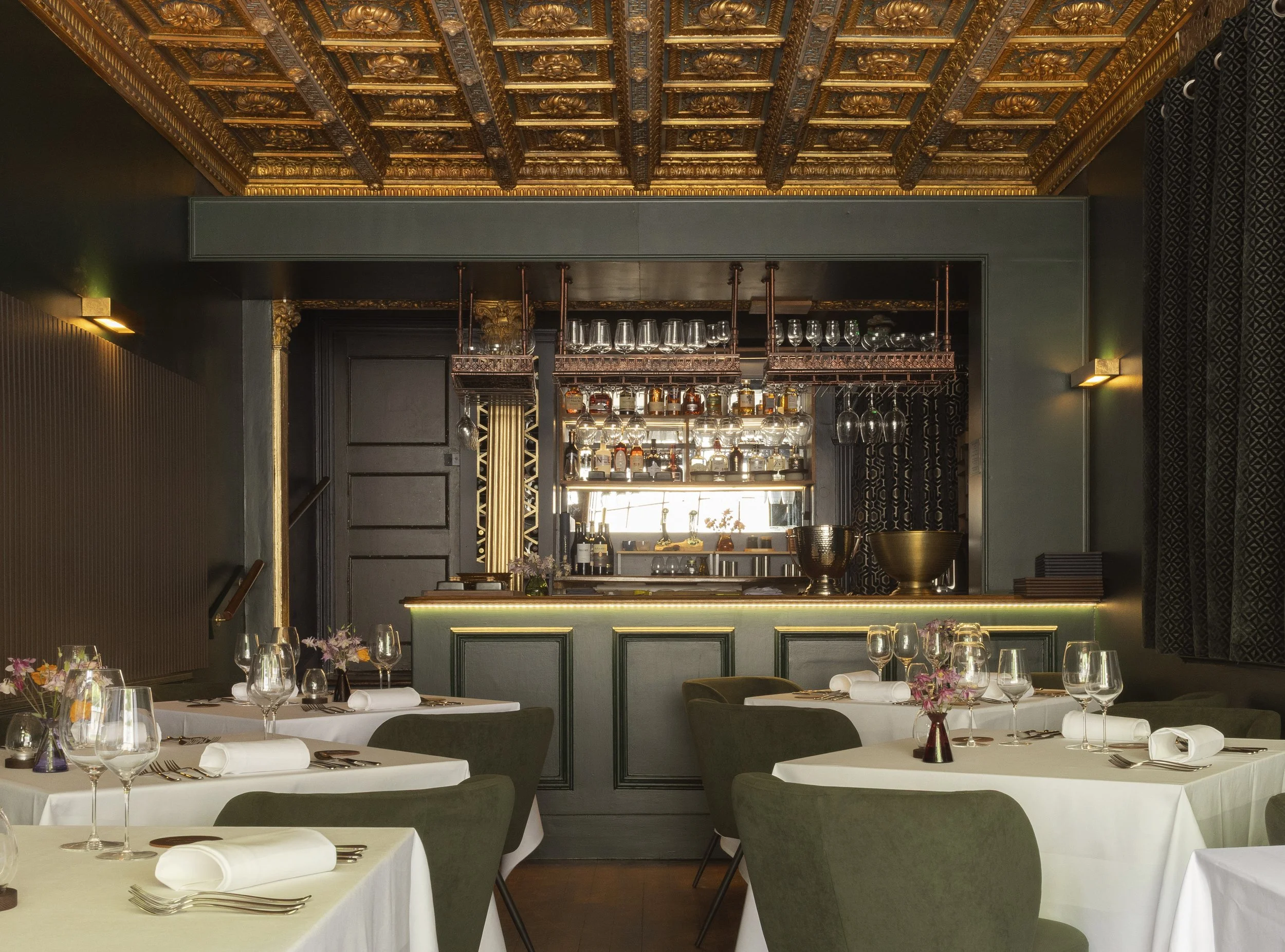 Elegant restaurant interior with white tablecloths, wine glasses, and simple floral centerpieces. Dark green chairs surround the tables. The background features a bar with hanging glasses and bottles, ornate gold detailing on the ceiling, black walls, and soft lighting.