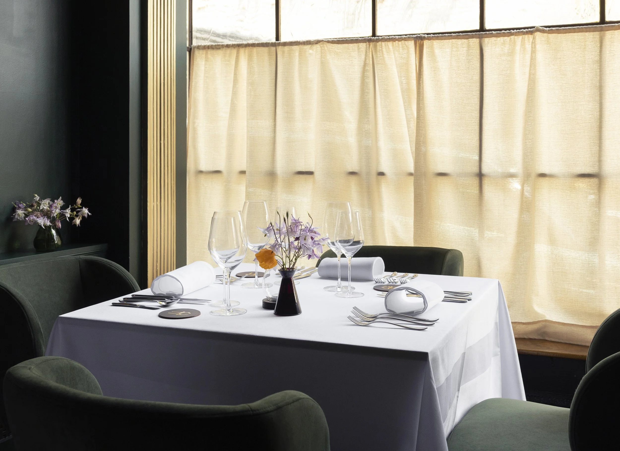 A dining table set with wine glasses, plates, silverware, and a floral centerpiece in a room with large windows and beige curtains.