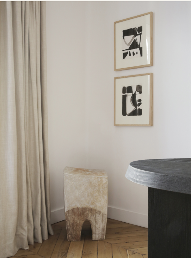 Corner of a room with a window and curtain, two abstract black and white framed artworks on the wall, a beige upholstered stool, and part of a dark wood table.