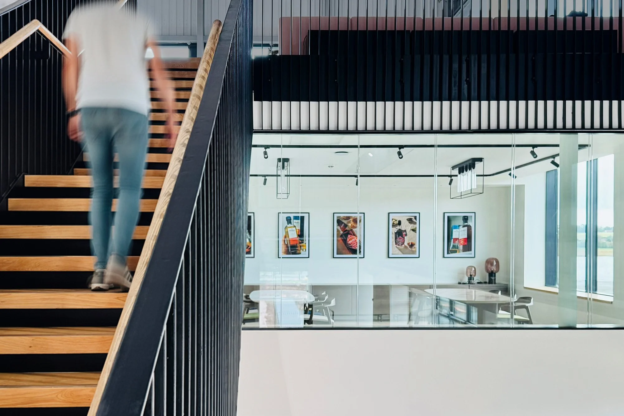 To the left of this image, a black metal and oak staircase rises to an upper room. To the right, we can see through a glazed screen into a room with artwork on the walls, and tables and chairs