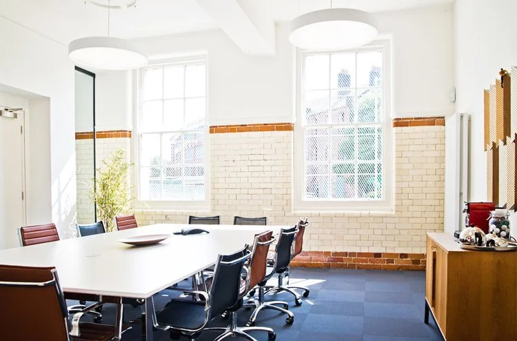 A bright meeting room with large windows, a white rectangular table surrounded by black and brown chairs, and a wooden cabinet with material samples on top.