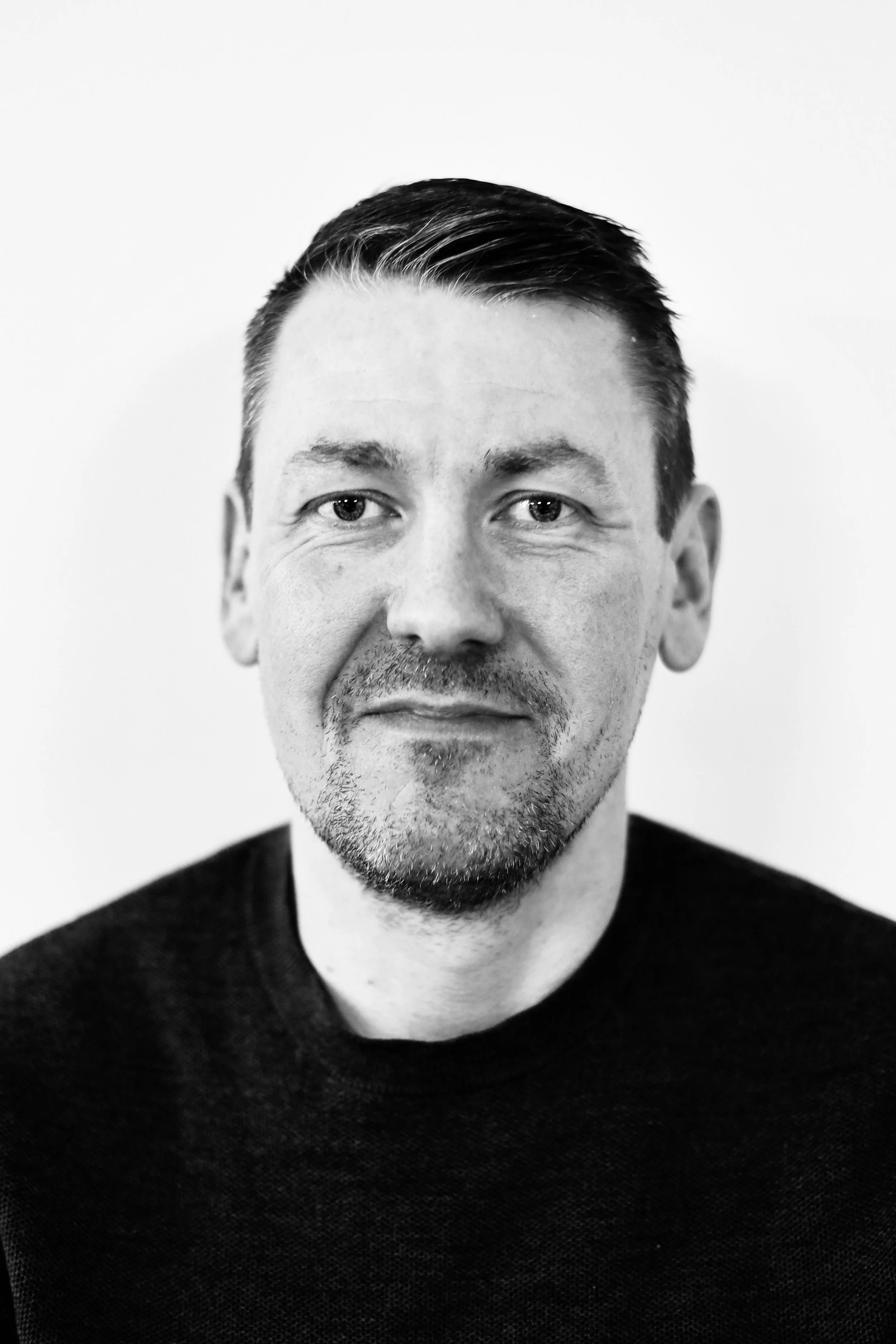A black and white portrait photograph of a man in his 40s. He is wearing a black t-shirt, has short cropped hair and is smiling at the camera