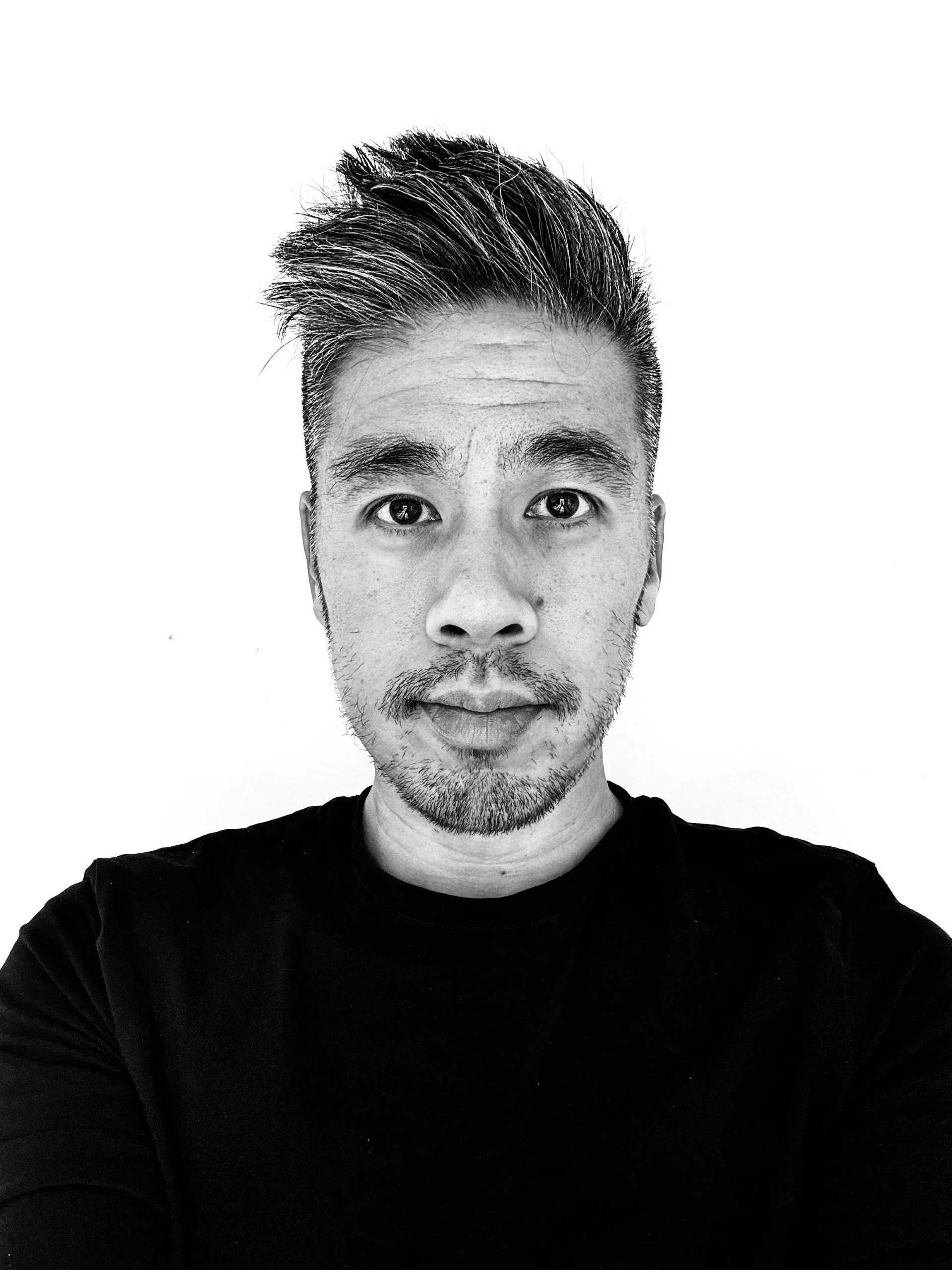 A black and white photograph of a man in his 40s. He is wearing a black t-shirt and has a quiff of hair