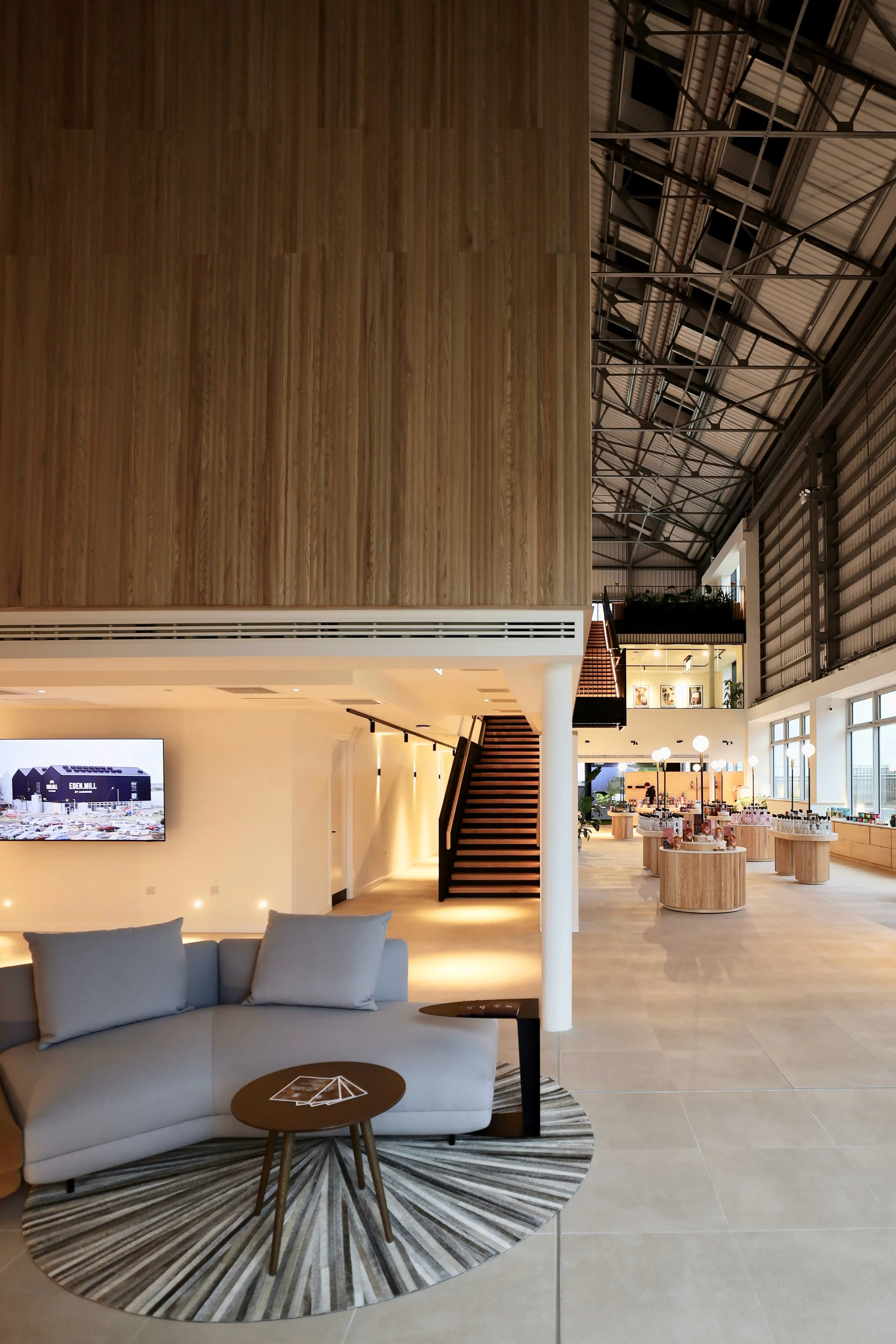 A bright, voluminous interior space with a black metal and oak feature stair rising to connect two upper floor levels. In the foreground is a grey, contemporary sofa and side tables