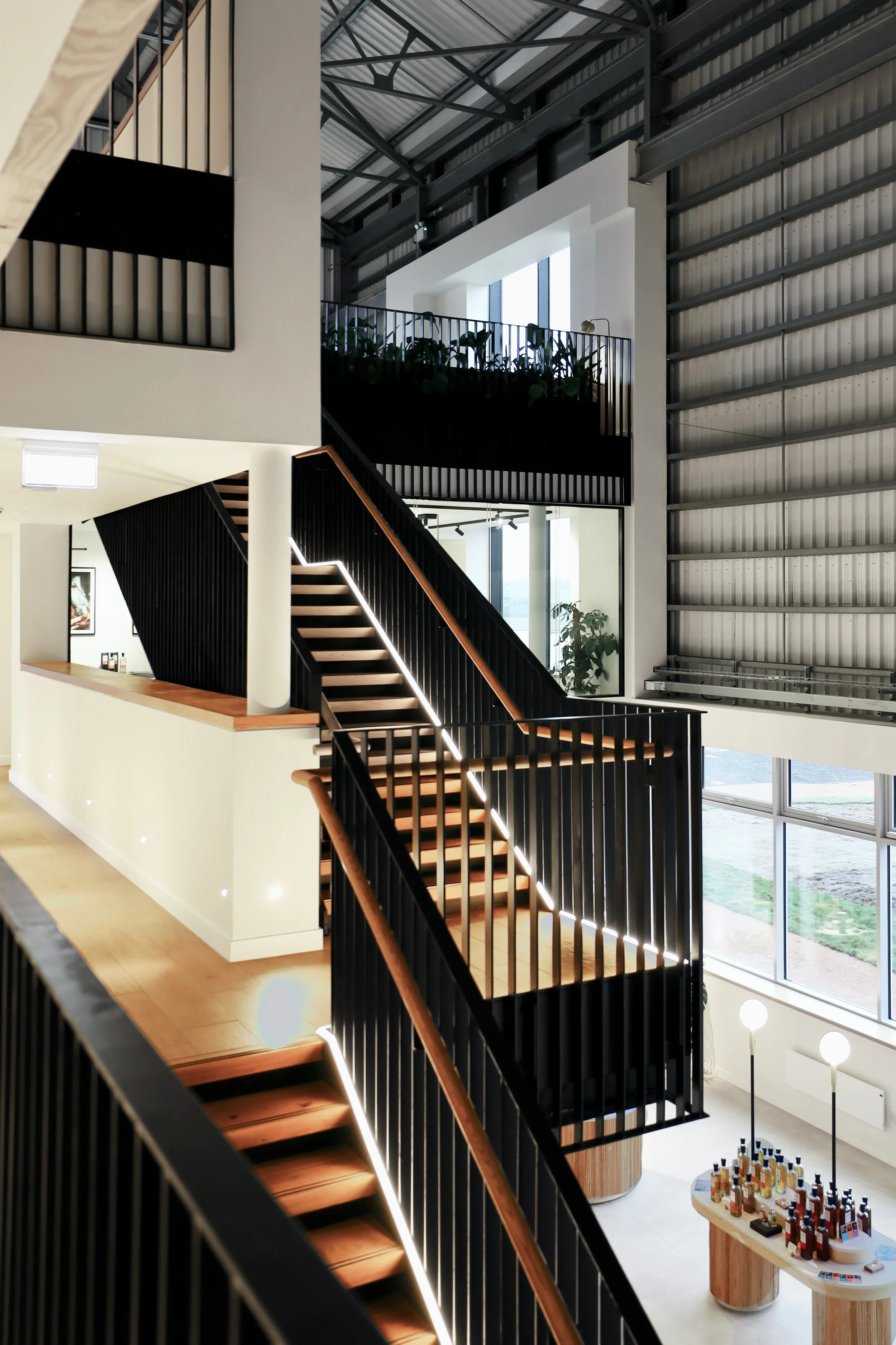 A black metal and oak staircase rises through the image, projecting out into the main atrium space of the distillery