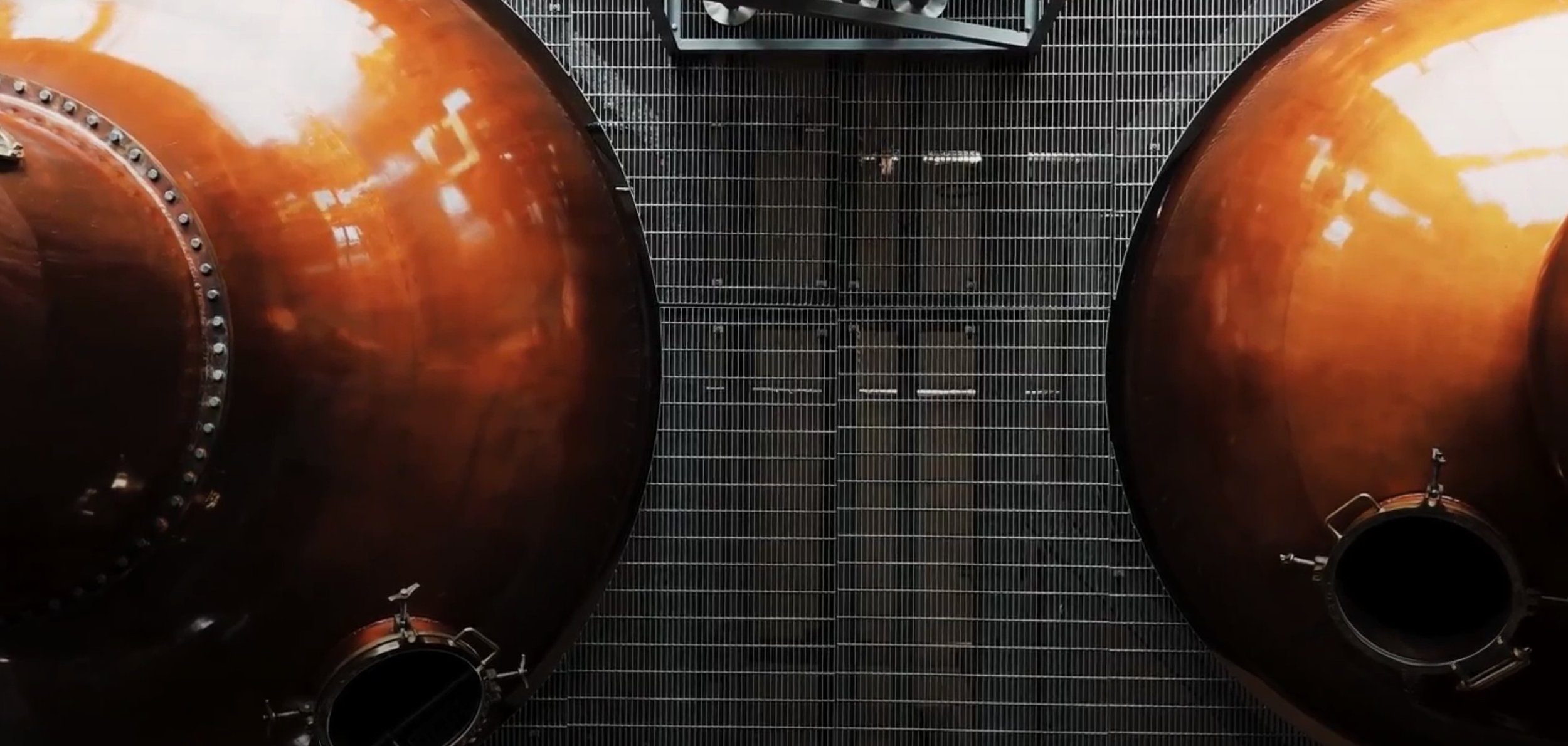 A top-down view of two polished copper whisky stills. There is steel grating between them.