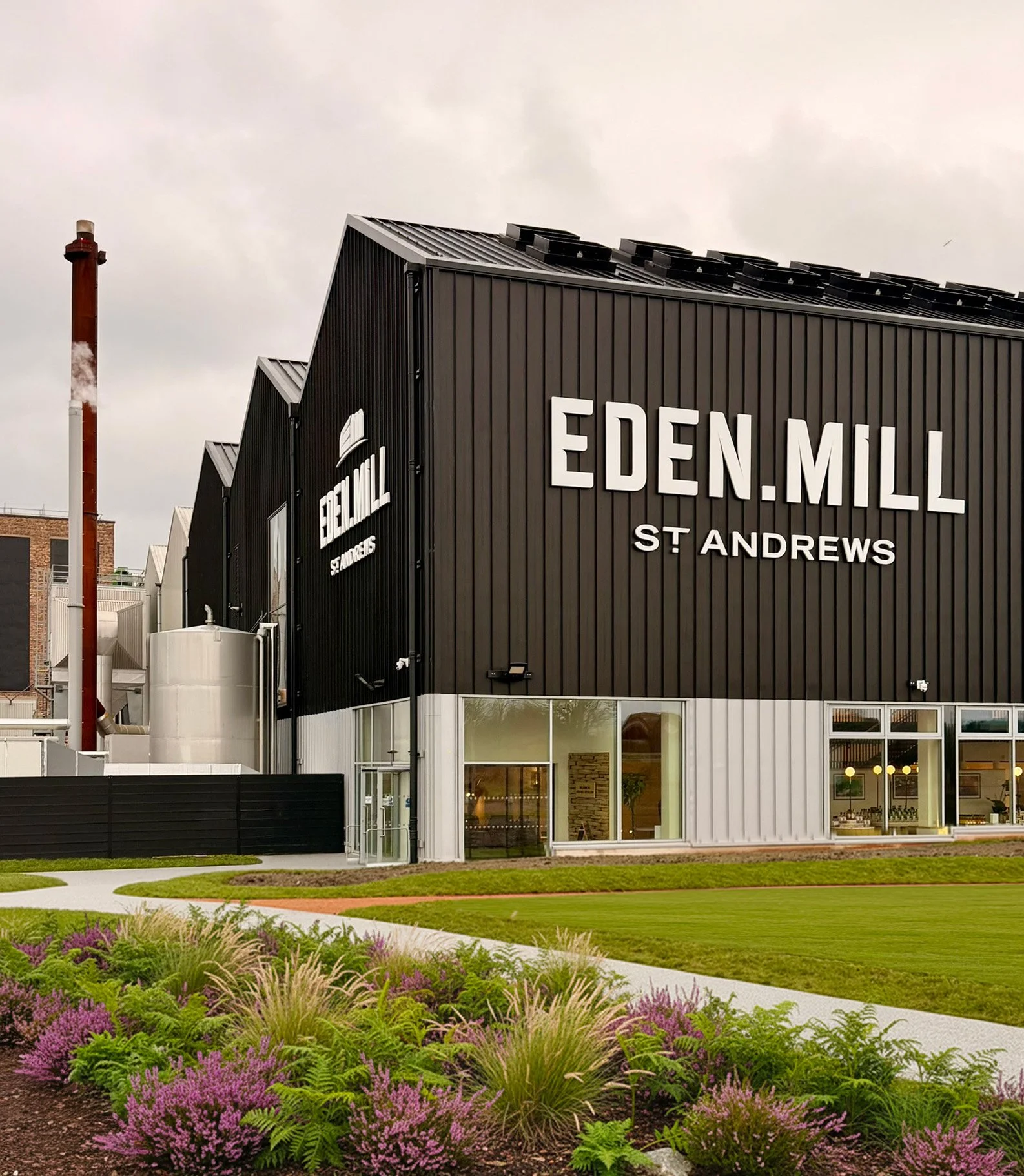 A tall black metal building with a pitched roof, surrounded by grass and planting. The words 'Eden Mill' are written on the side, in large white letters