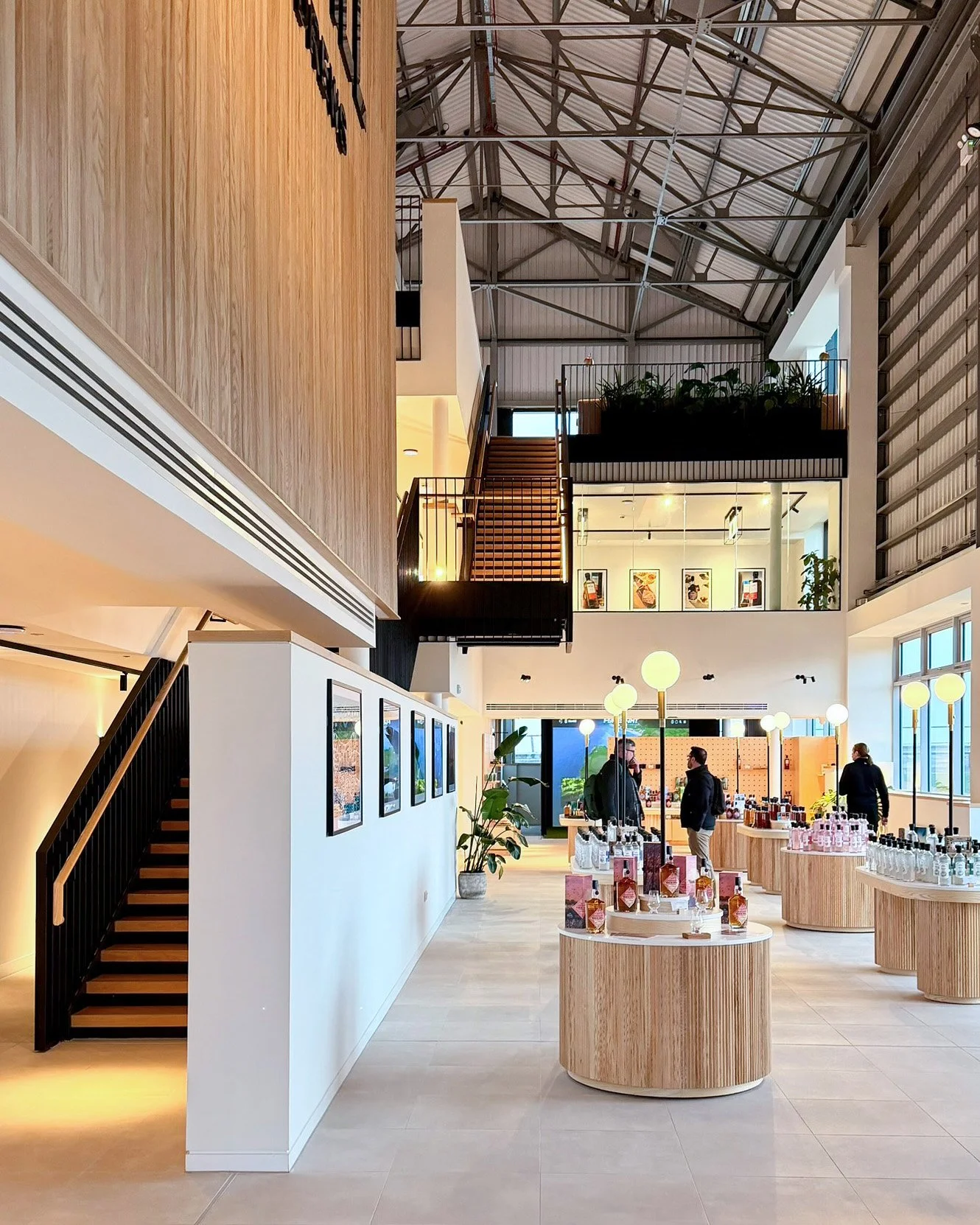A bright, voluminous interior space with a black metal and oak feature stair rising to connect two upper floor levels. On the ground floor, there are shop displays of whisky and gin products