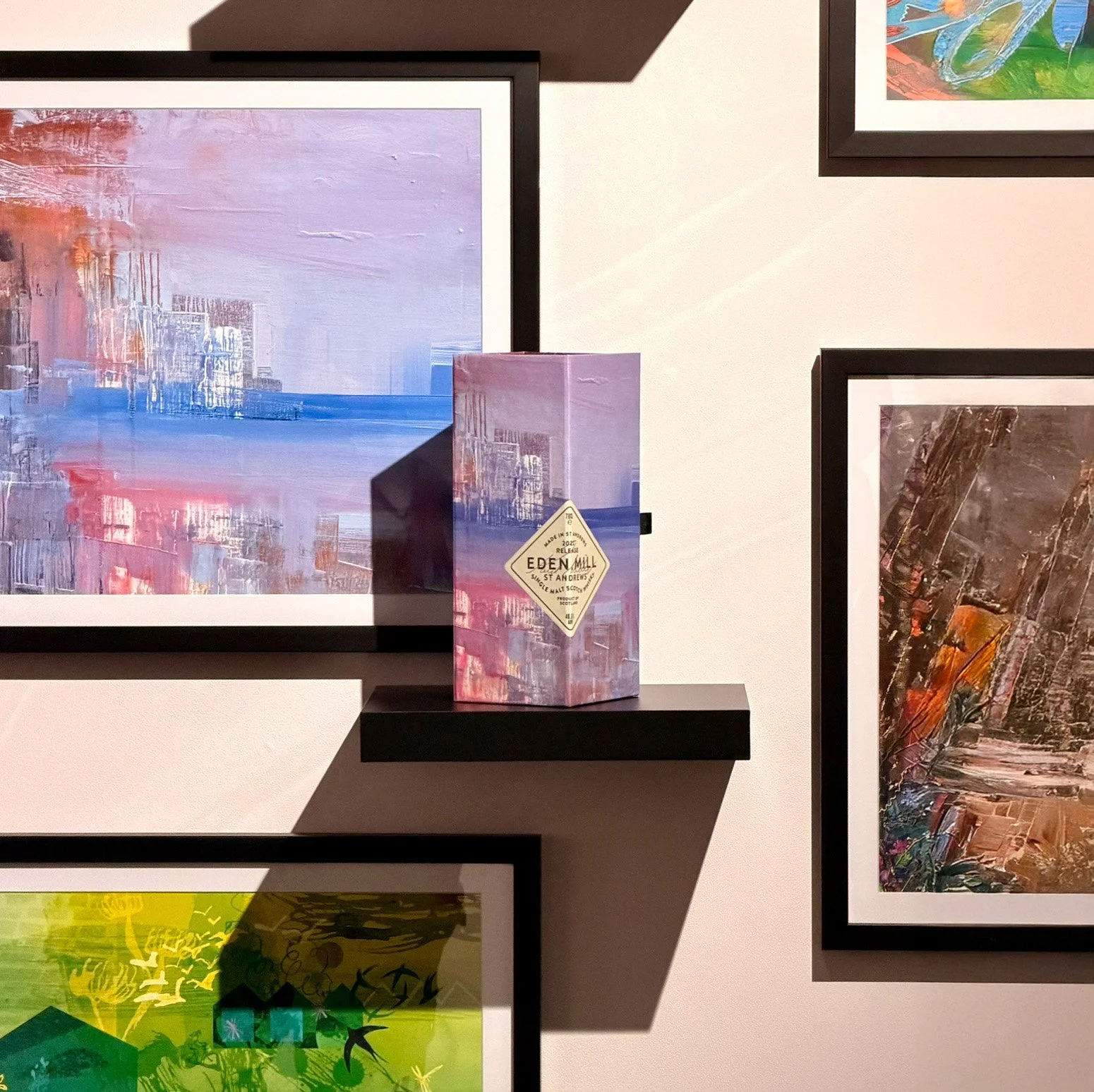 An artfully decorated Eden Mill whisky box is displayed on a floating shelf, surrounded by framed prints of abstract paintings