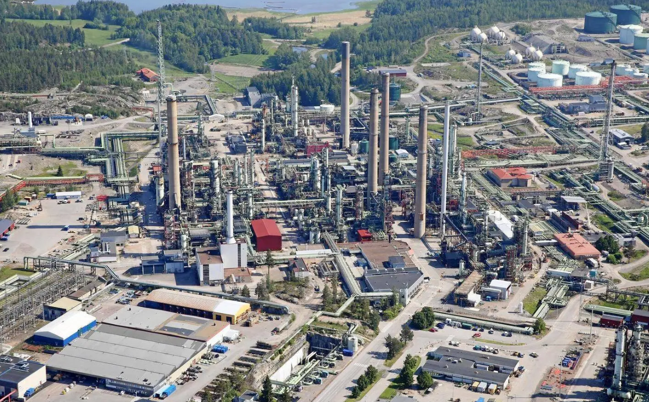 Kuva teollisuuslaitoksesta, jossa on paljon putkia, torneja ja varastoja, ympäröitynä metsäisillä alueilla ja rakennuksilla.