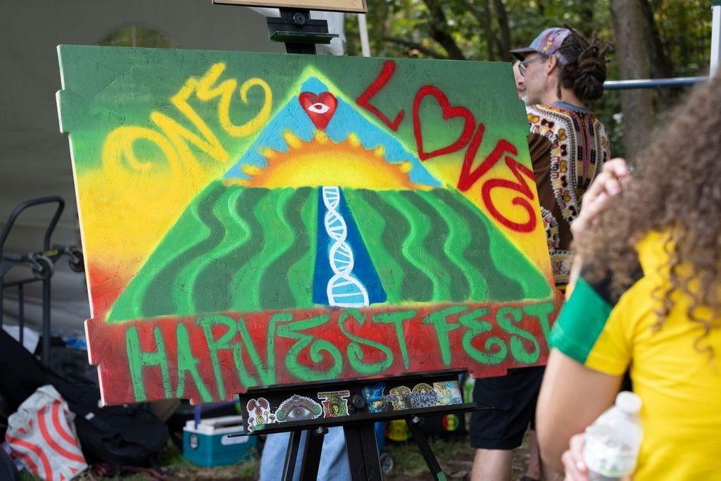 Colorful poster at an outdoor event depicts a stylized agricultural scene with a mountain, sun, and a river of DNA strand, with words "ONE LOVE HARVEST FEST" in bold letters, celebrating unity and harvest.