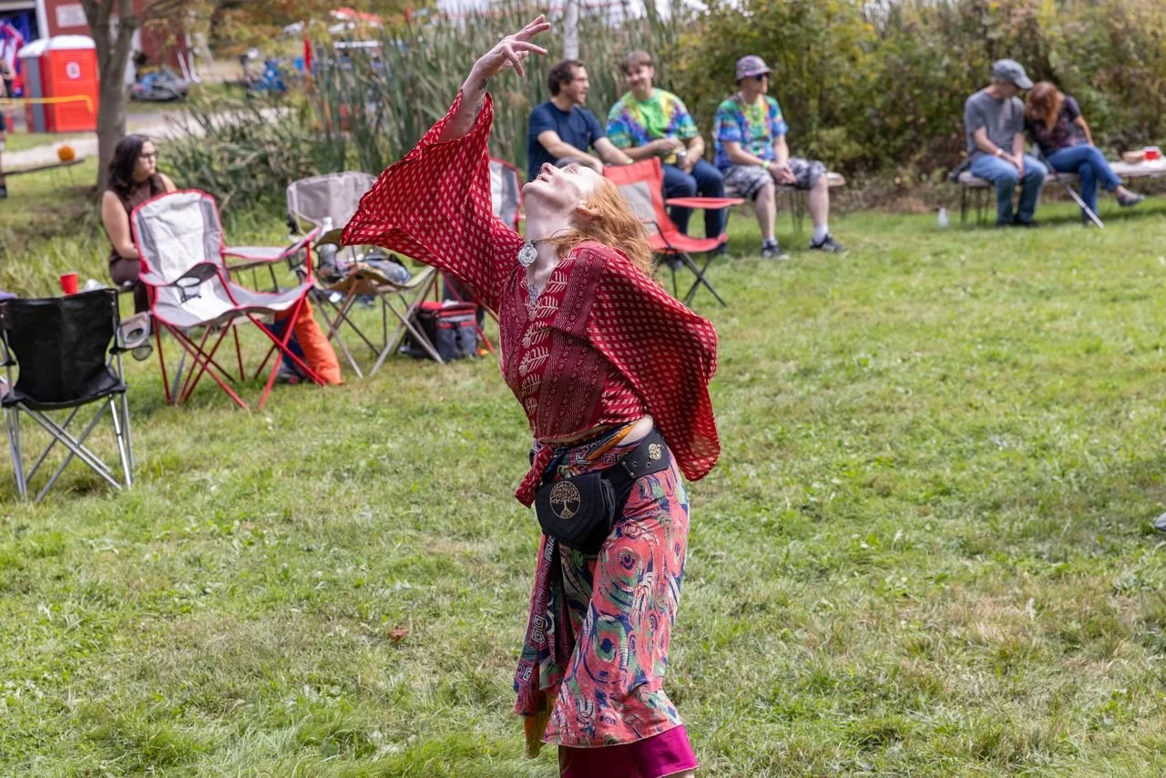 A woman in colorful, patterned pants and a red shawl dances in a grassy outdoor area during a gathering, while people in the background sit on lawn chairs watching.