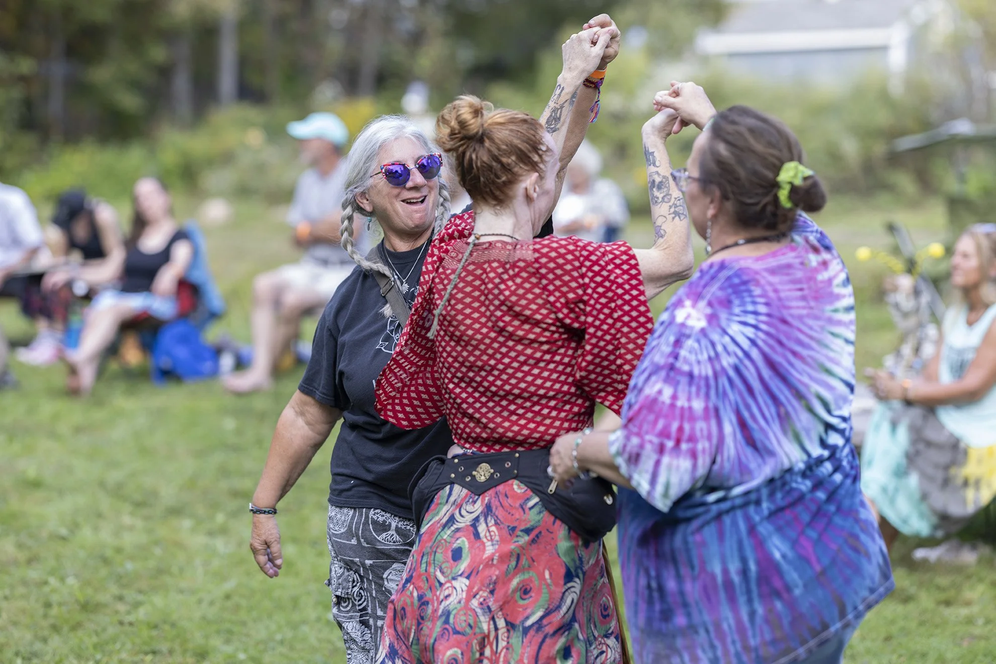 Three women dancing together outdoors at a gathering, with others seated on the grass in the background.