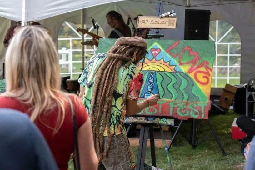 A woman with dreadlocks painting a colorful artwork at an outdoor festival, with the words 'Love' and 'Fest' visible, under a white canopy tent, with a small audience watching.