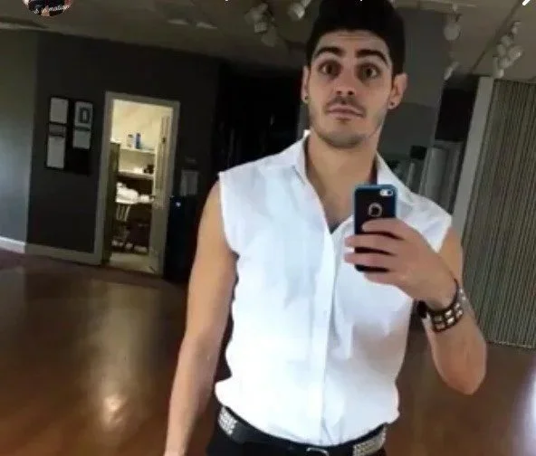 A young man taking a mirror selfie in a room with dark walls and wooden floors, wearing a sleeveless white shirt and black pants.