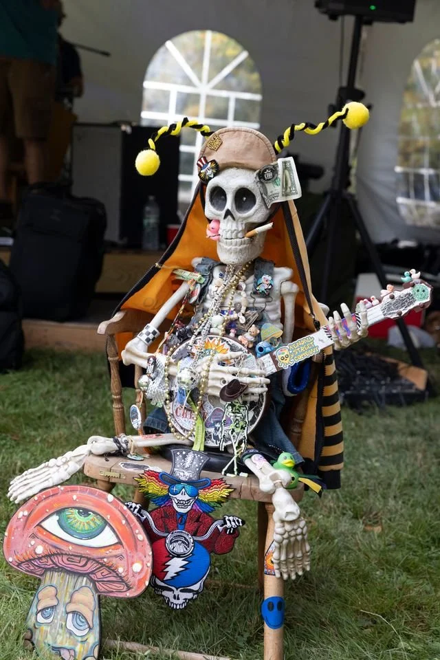 A decorative skeleton seated on a wooden chair coated with accessories, holding a guitar, with a large eye and a clown sticker on the ground in front. The skeleton is adorned with jewelry, a candy cigarette, dollar bills, and wears a hat with yellow 