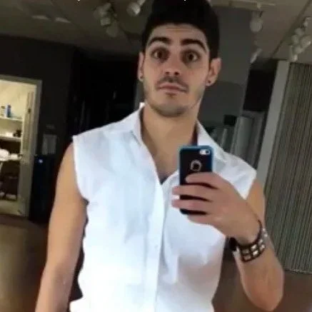 Young man taking a selfie in a mirror, wearing a sleeveless white shirt and wristbands.