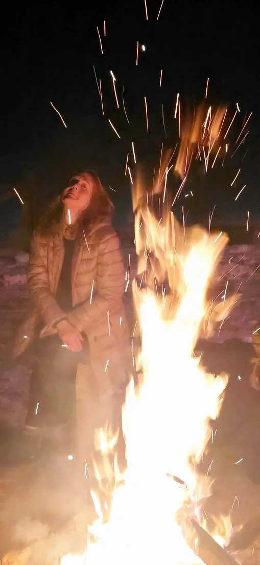 Person sitting by a campfire at night, smiling and looking up as sparks fly upward.