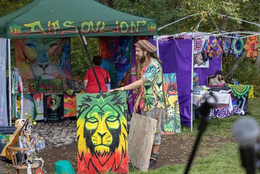 An outdoor market stall selling tie-dye and psychedelic art, with a man wearing a patterned shirt and hat holding a lion-themed painting, and other stalls selling more colorful artwork and accessories.