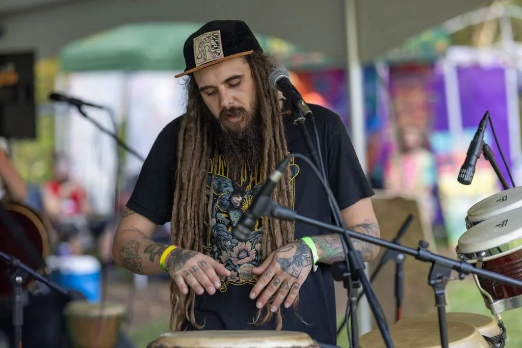 A musician with long dreadlocks, tattoos, and a black cap playing percussion instruments at an outdoor event.