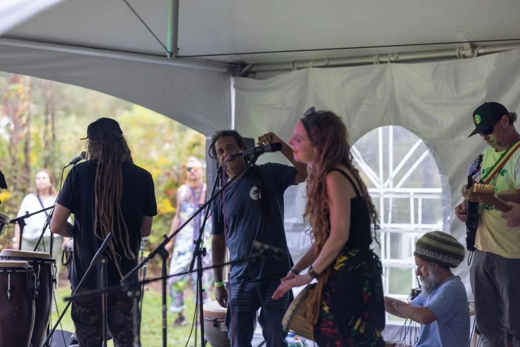 A group of musicians performing on a stage under a white tent, with outdoor scenery visible in the background.
