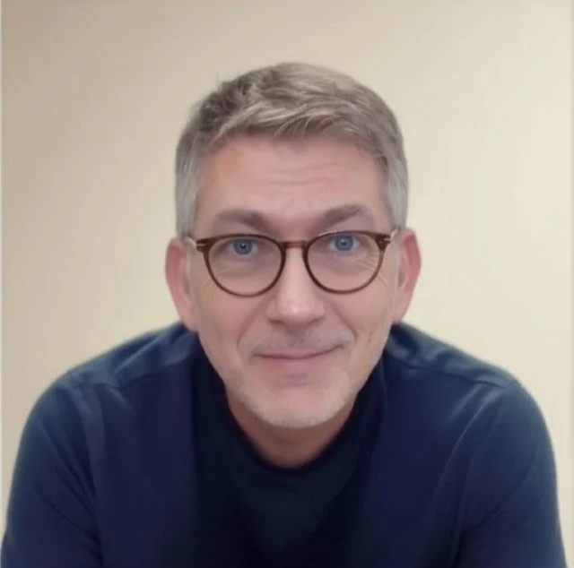 A middle-aged man with short gray hair, glasses, and a slight smile, wearing a dark shirt, sitting in front of a plain light-colored wall.