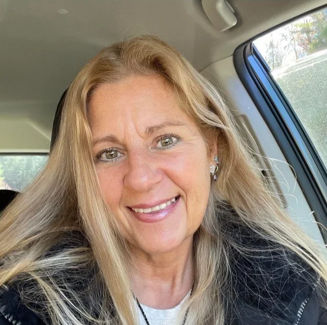 A woman with blonde hair and blue eyes smiling inside a vehicle, with the car interior and window visible in the background.