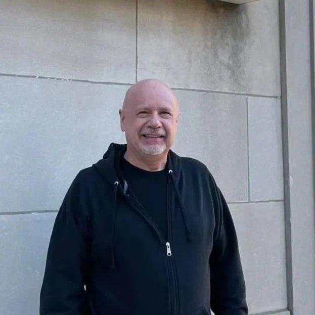 A smiling bald man with a goatee wearing a black hoodie and shirt standing in front of a light gray stone wall.