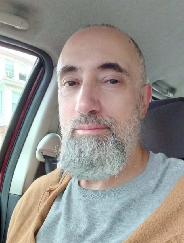 A middle-aged man with a beard and short hair sitting inside a vehicle, smiling slightly at the camera.