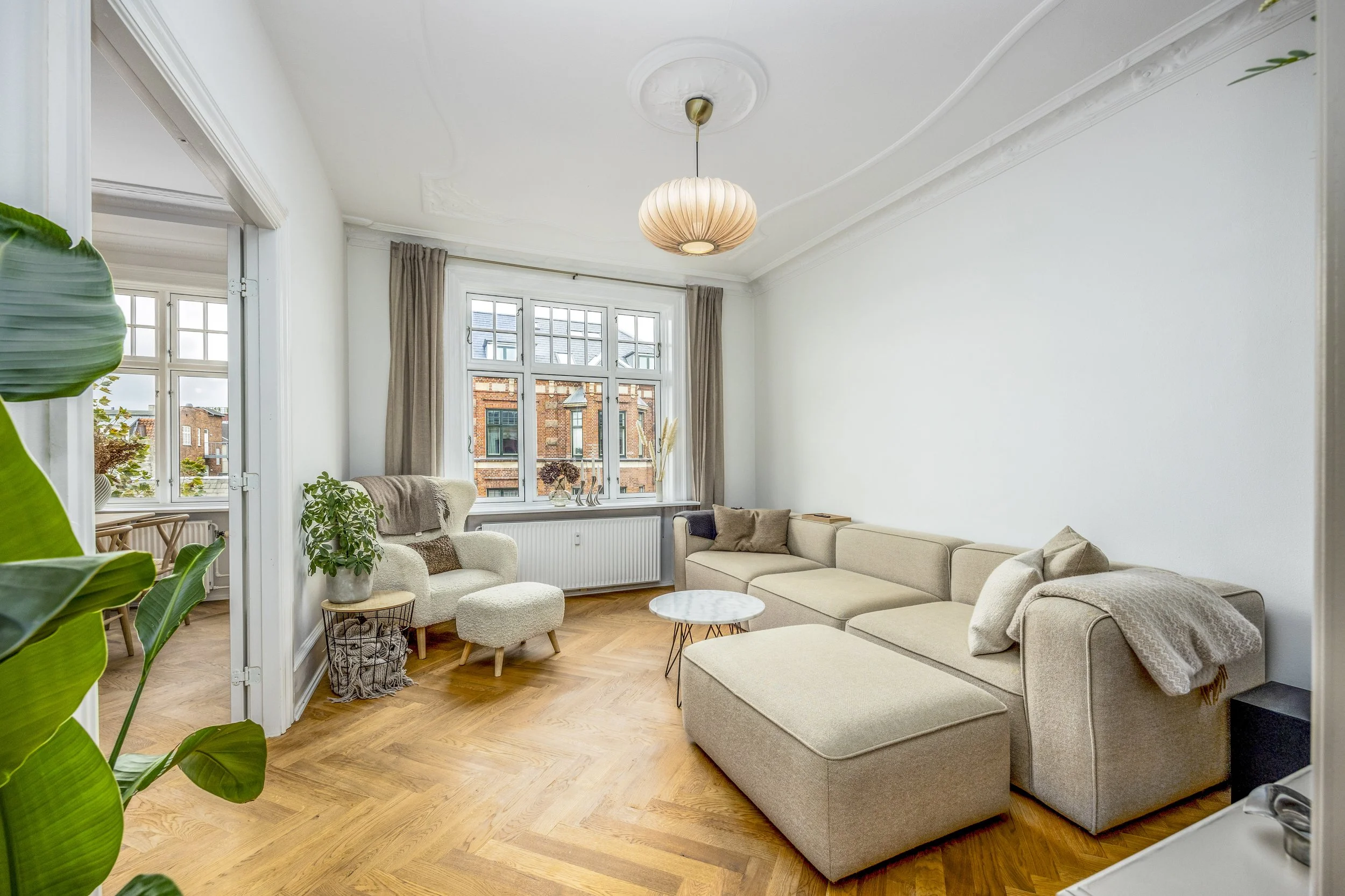 A living room with a large cream-colored sectional sofa, a matching armchair with an ottoman, a small round coffee table, and large windows with beige curtains. The room has hardwood floors and decorative ceiling moldings, with city buildings visible outside the windows.