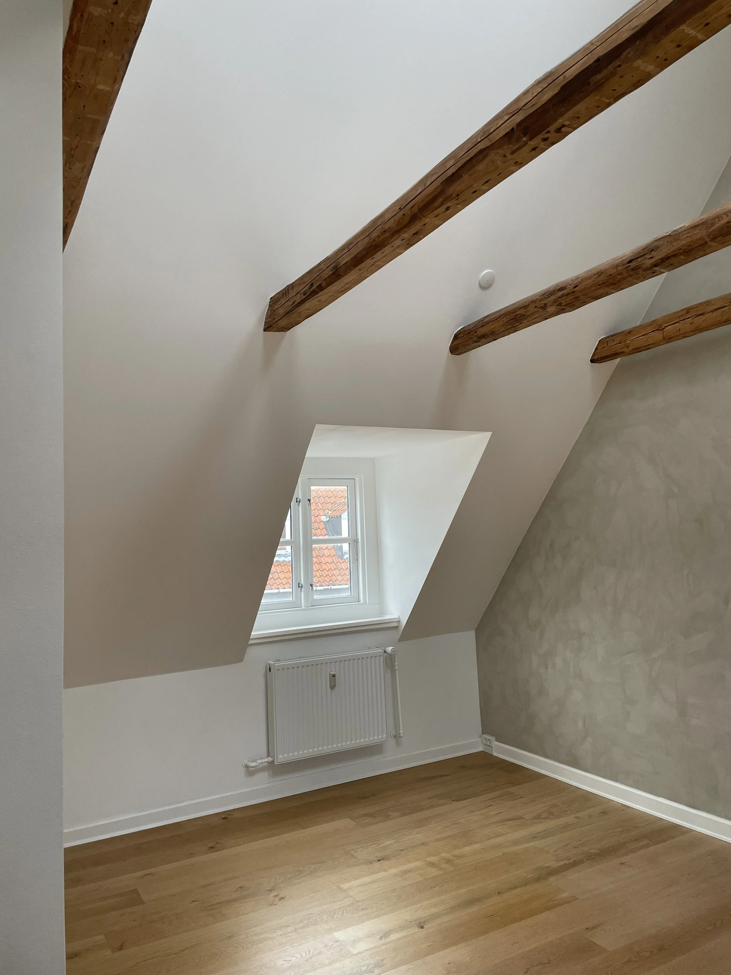 Empty attic room with white walls, a wooden floor, exposed wooden beams on the ceiling, a small window with a view of red tile roofs, and a radiator below the window.