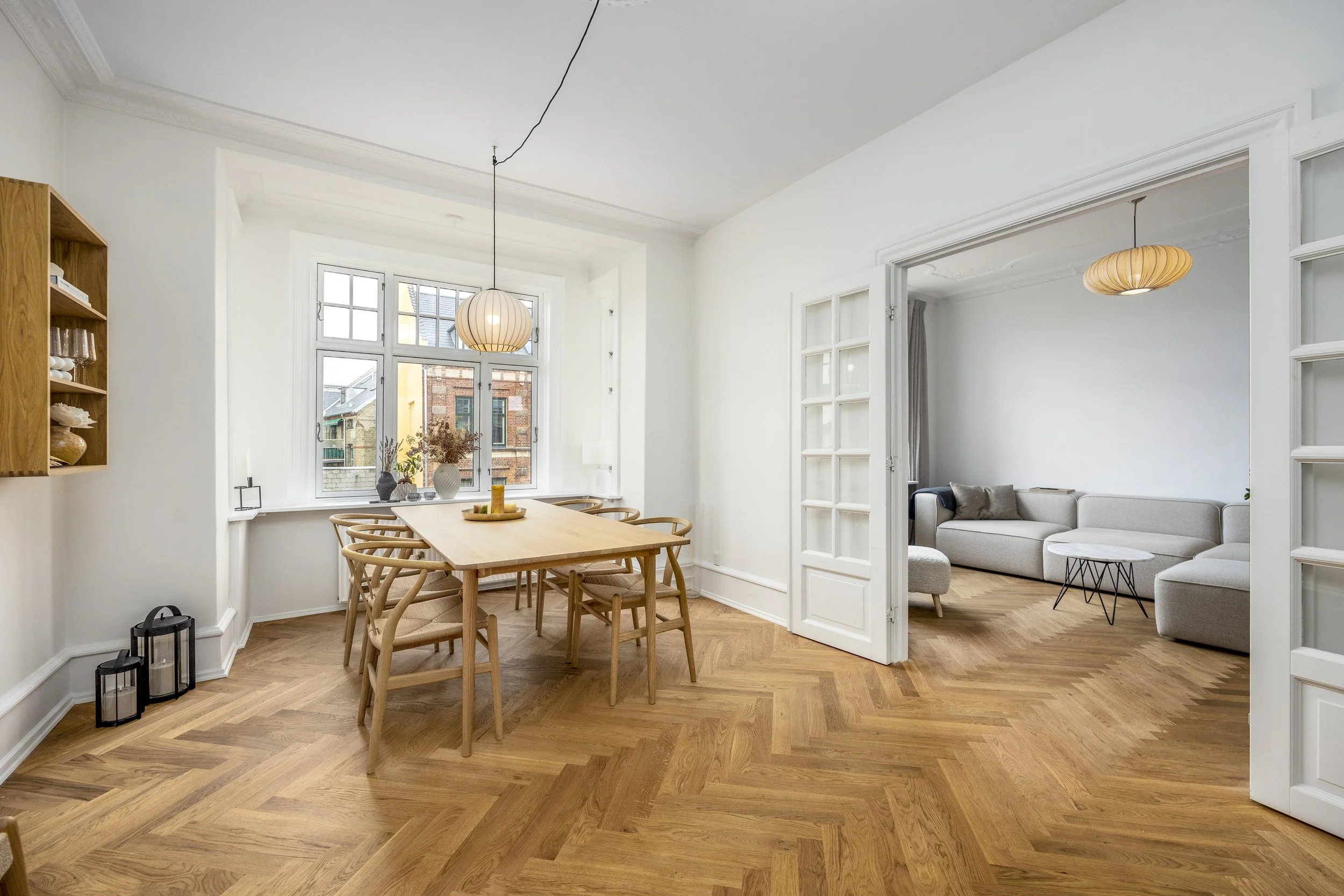 Dining area with a wooden table and six chairs, large window with a view of brick buildings, and a living room with a gray sectional sofa and round coffee table, all in a bright, modern decor.