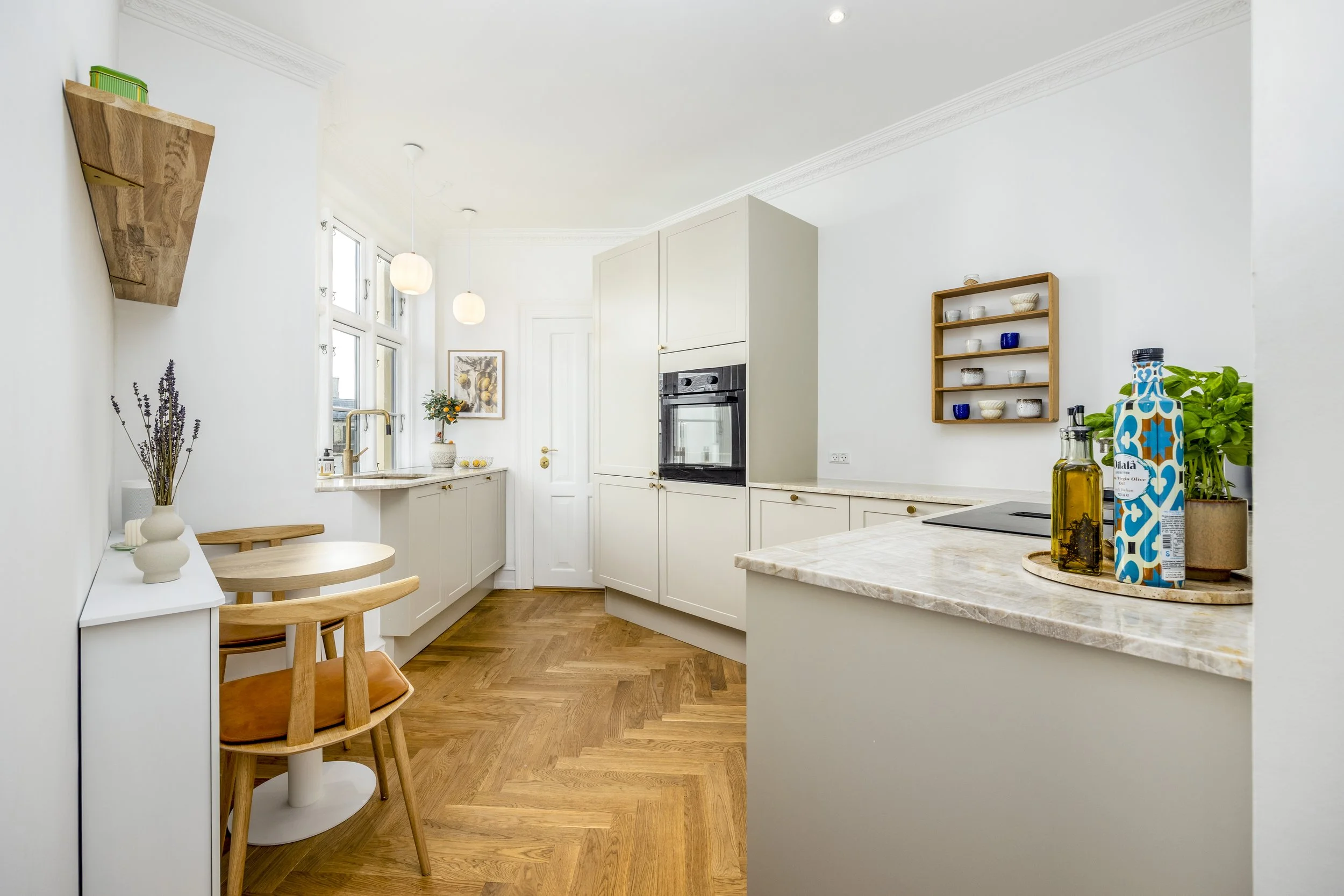 Bright modern kitchen with white cabinets, wood flooring, marble countertops, and natural light from large windows. Decor includes a small table with a chair, a wall-mounted wooden shelf with bowls, and bottles of oil and condiments on the counter.